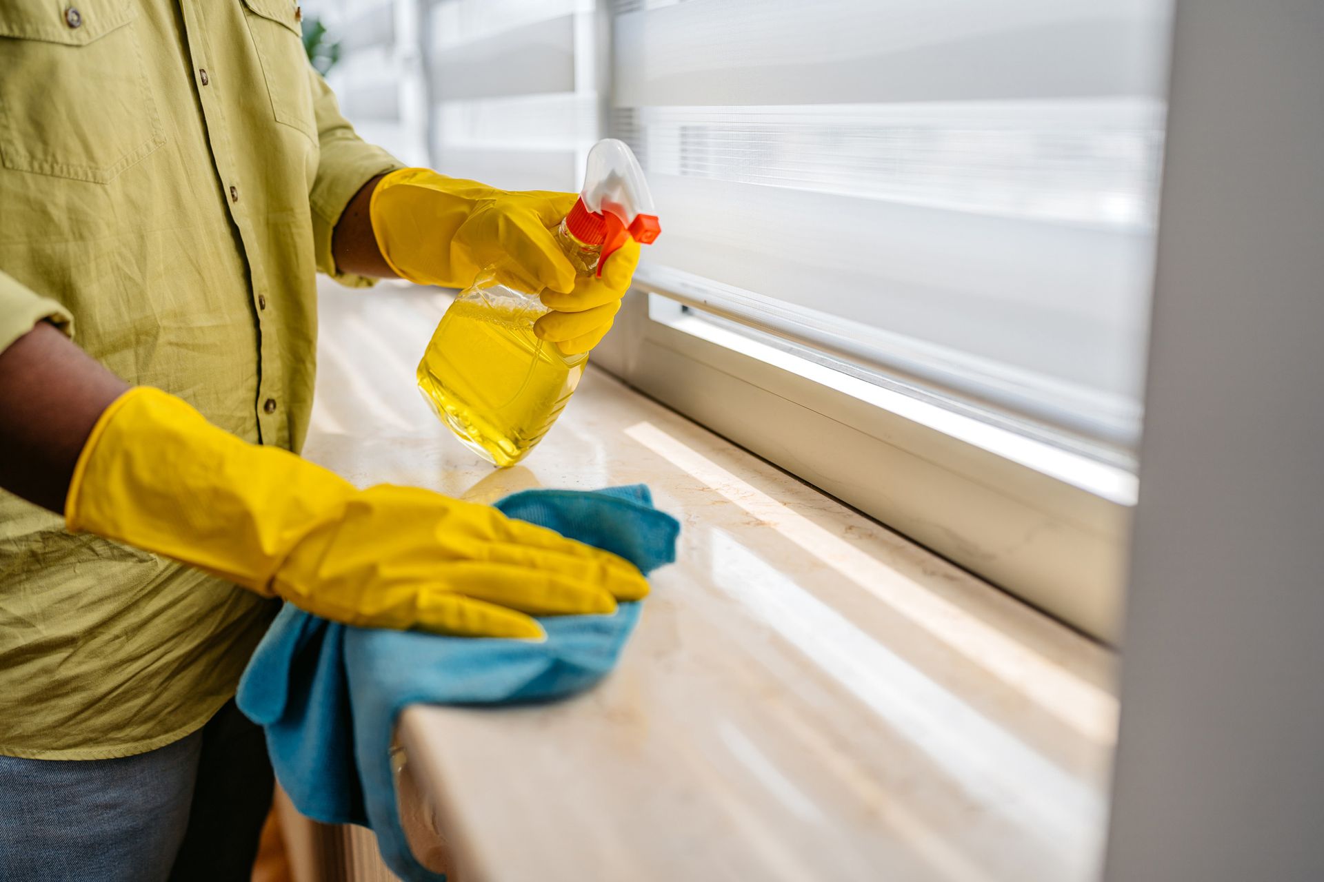 Person wearing yellow gloves spraying and wiping a windowsill with a blue cloth.