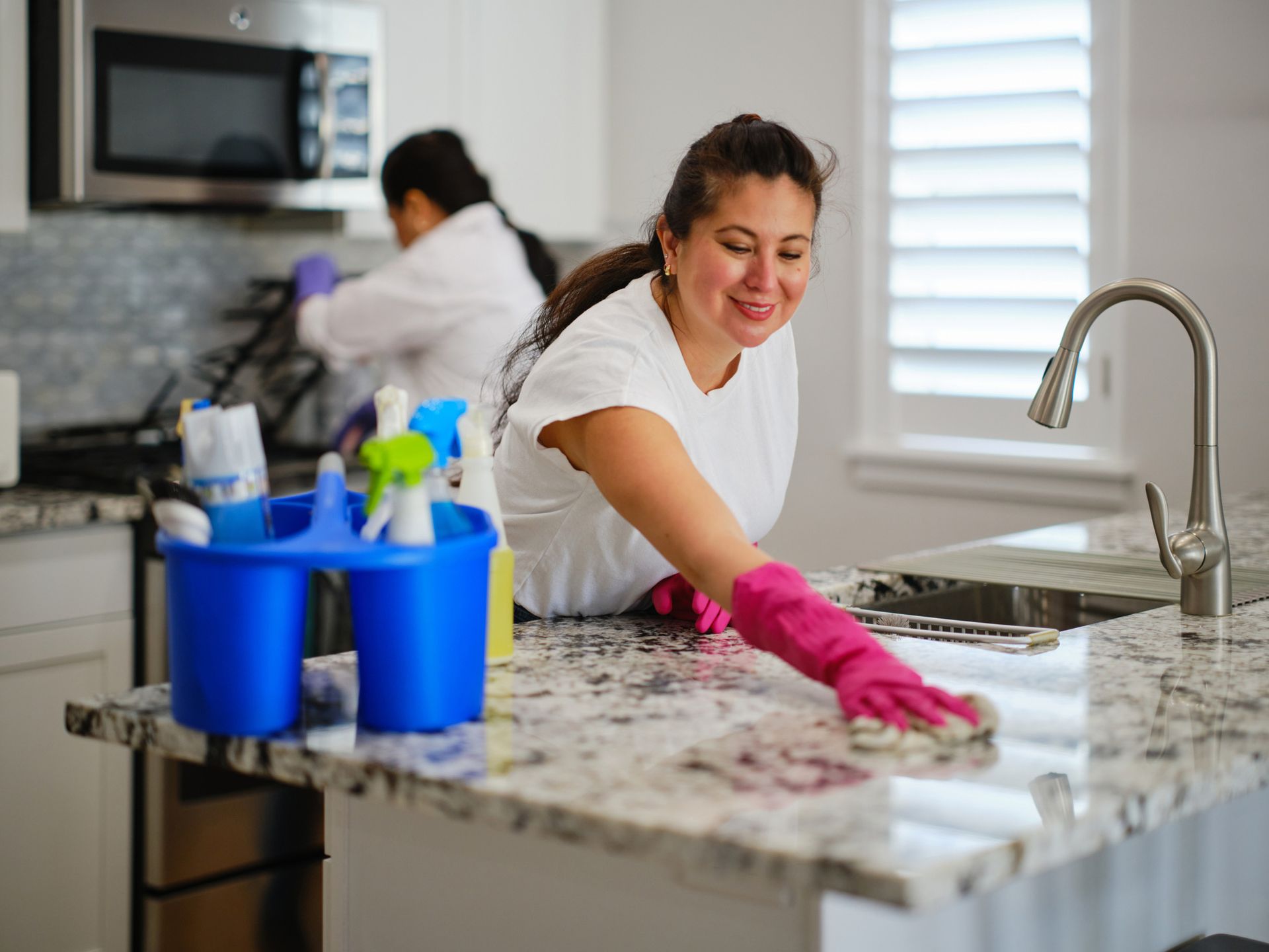 Two women cleaning a kitchen. One wipes a countertop with a pink-gloved hand, smiling. 