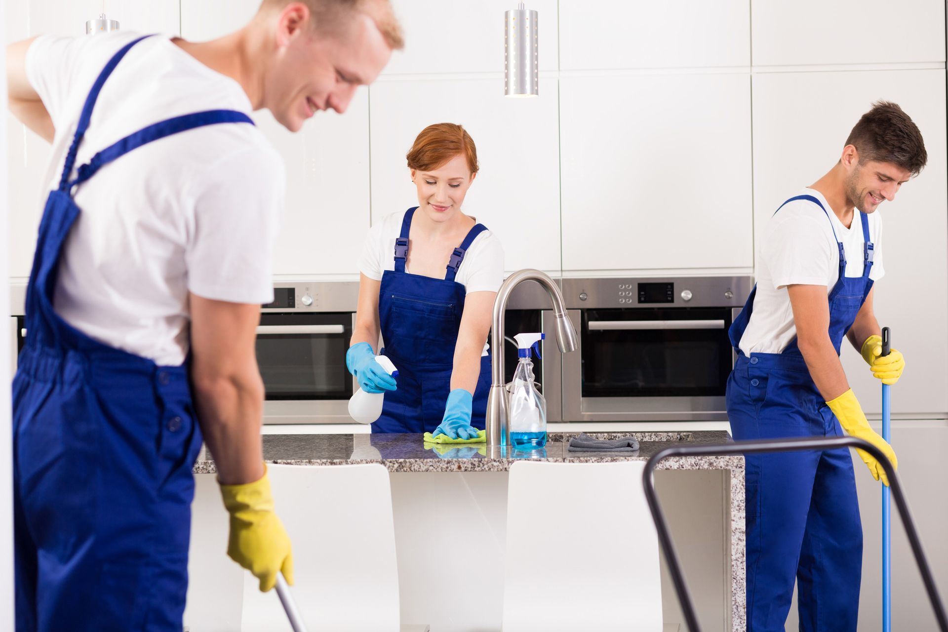 Three cleaning professionals, in blue overalls, cleaning a kitchen.
