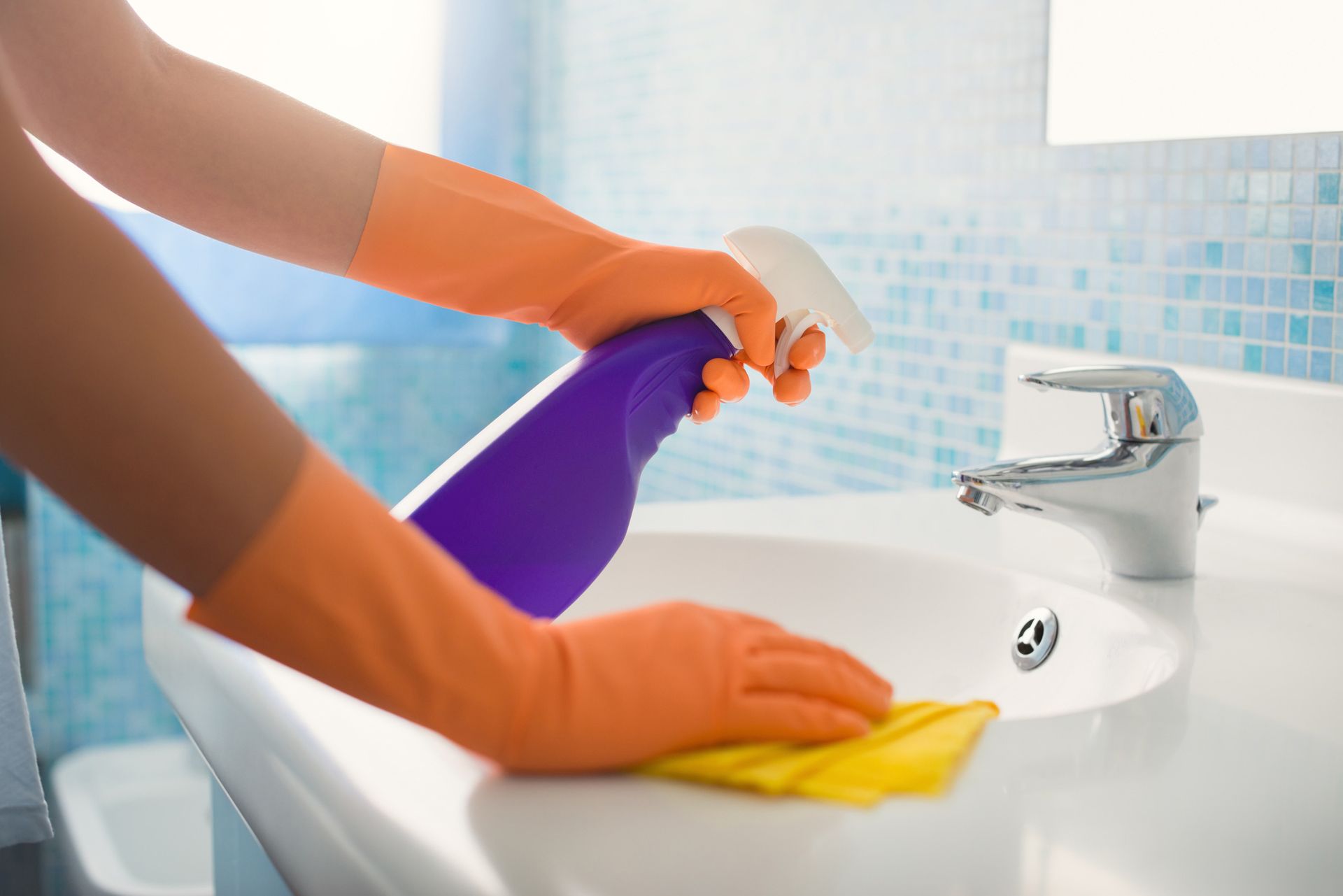 Hands in orange gloves spraying and wiping a white bathroom sink with a spray bottle and yellow cloth.