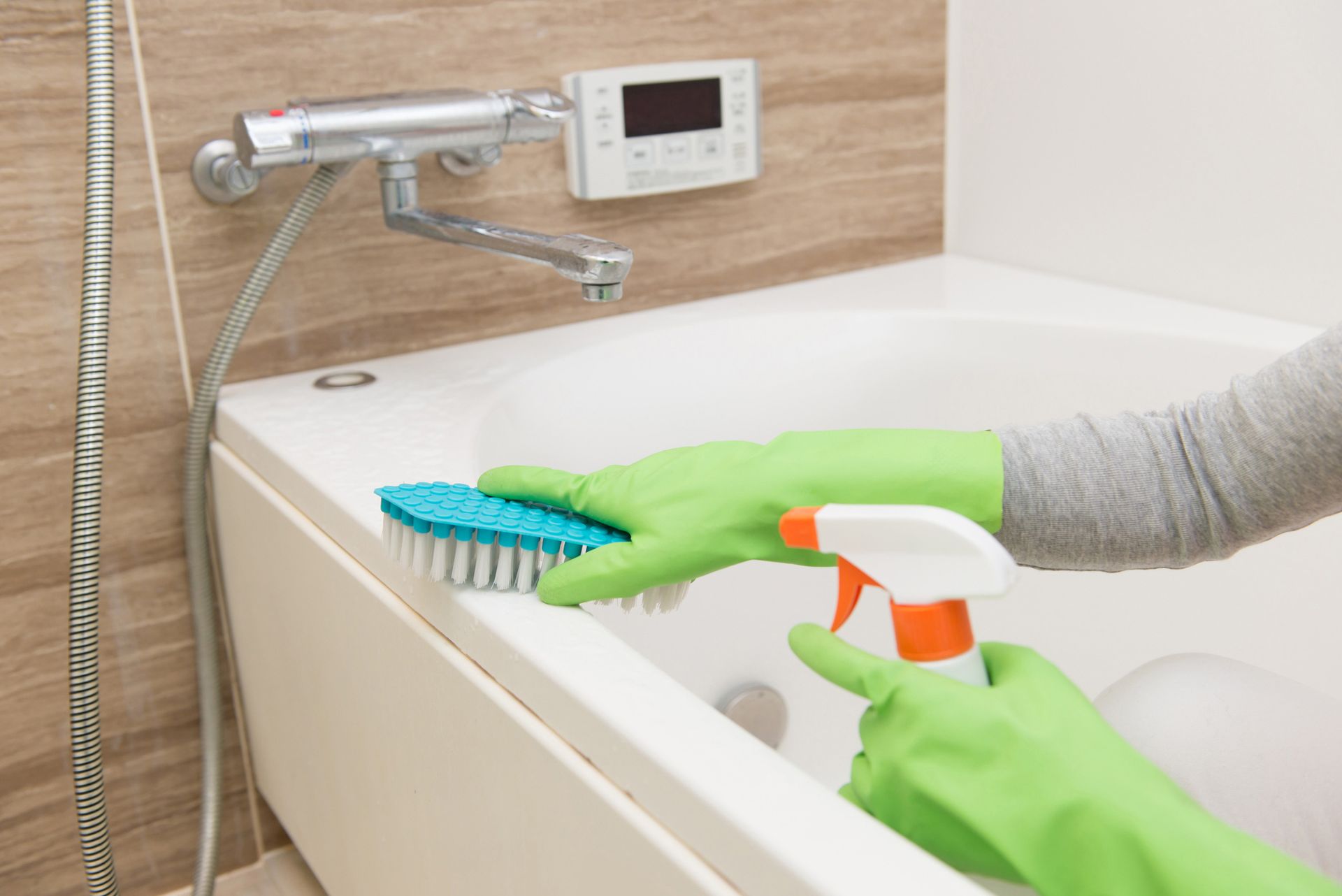 Person wearing green gloves cleaning a white bathtub with a scrub brush and spray bottle.
