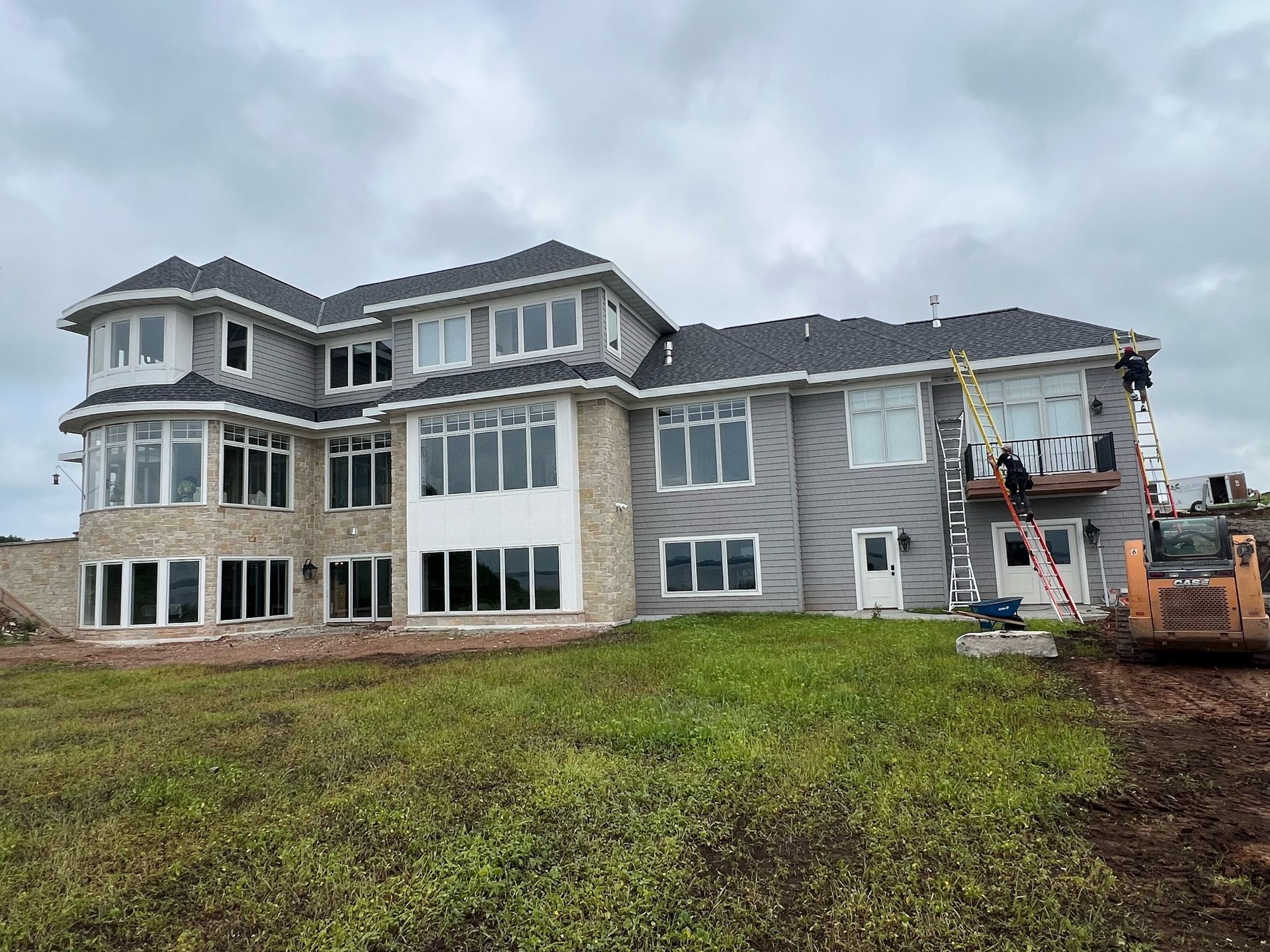 A two-story house under construction with grey siding and stone accents, surrounded by a grassy lot and a small bulldozer.
