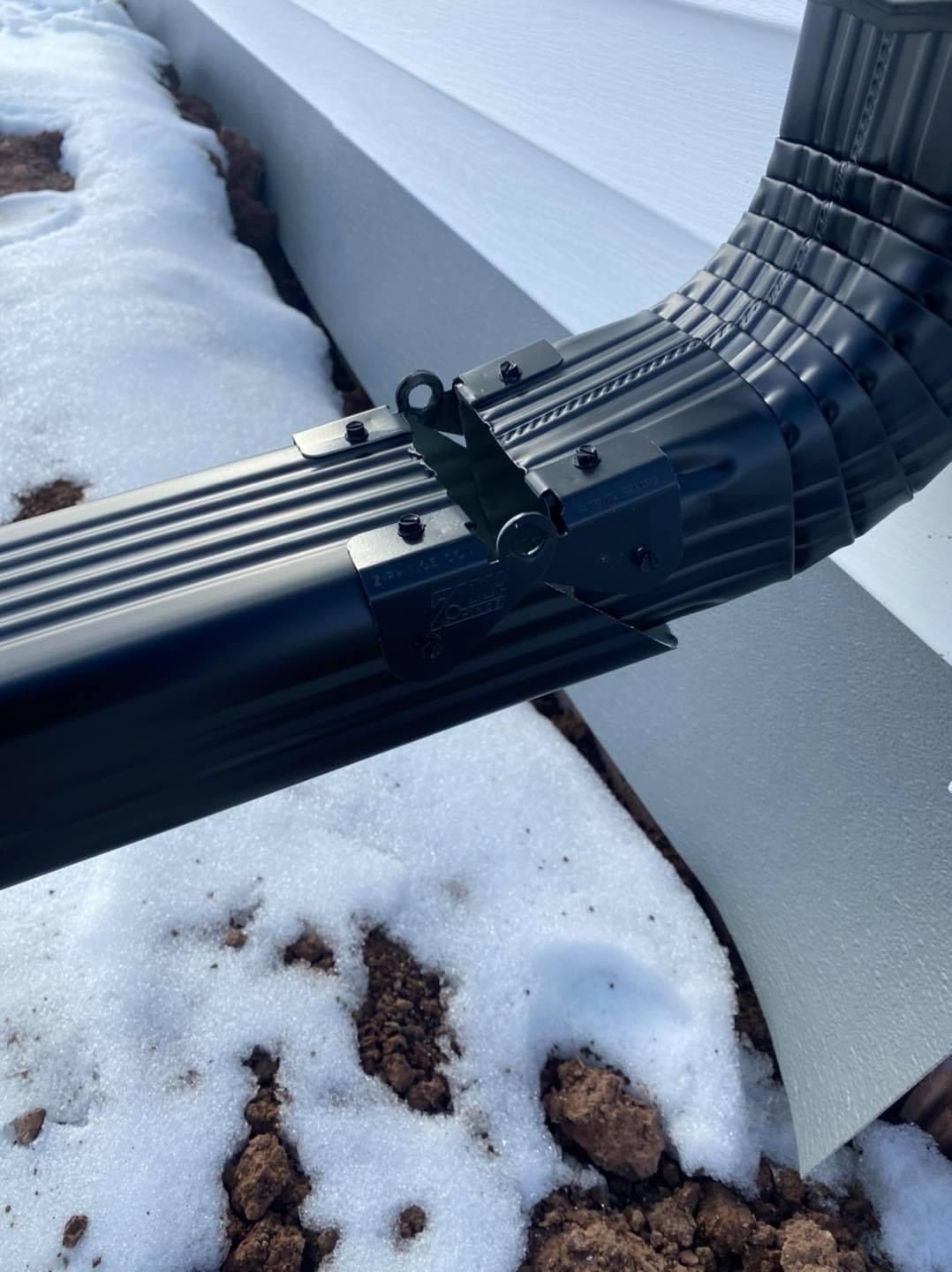 A close-up of a black gutter downspout hinge connecting a horizontal extension to a vertical elbow over snowy ground.