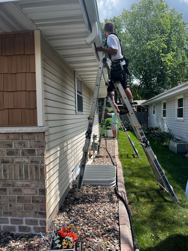A person wearing a utility belt stands on an A-frame ladder, working on the gutters of a tan home exterior.