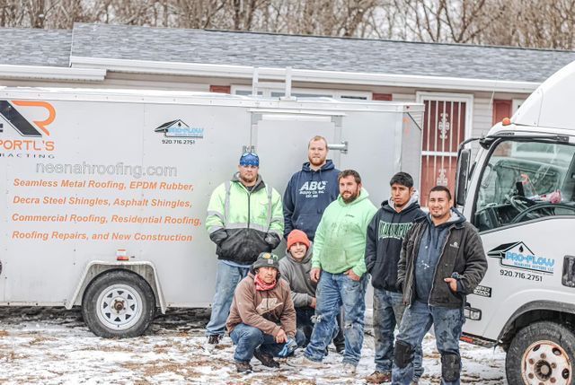 A group of workers in casual and work clothing stand outside near a roofing company trailer in a snow-dusted landscape.