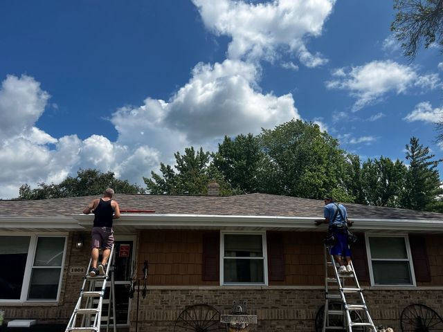 Two workers on ladders install gutters on the front of a brown, single-story house under a blue, cloudy sky.