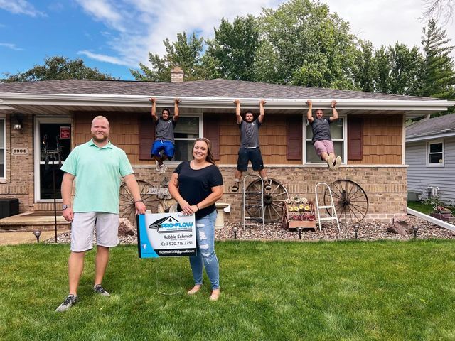 A man and woman stand in a lawn holding a sign, while three others hang from the house's rain gutters.