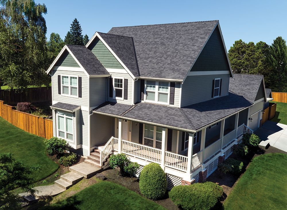 A two-story grey house with a wraparound porch, green gables, and a dark shingled roof, surrounded by a grassy lawn.