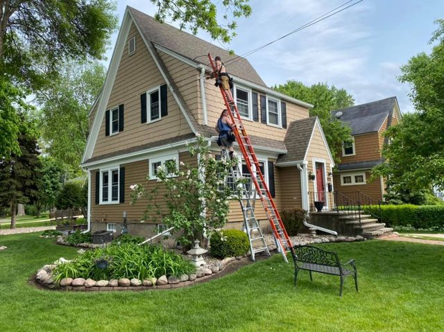 A person on a tall ladder cleans the gutter of a light brown, two-story house with black shutters on a sunny day.
