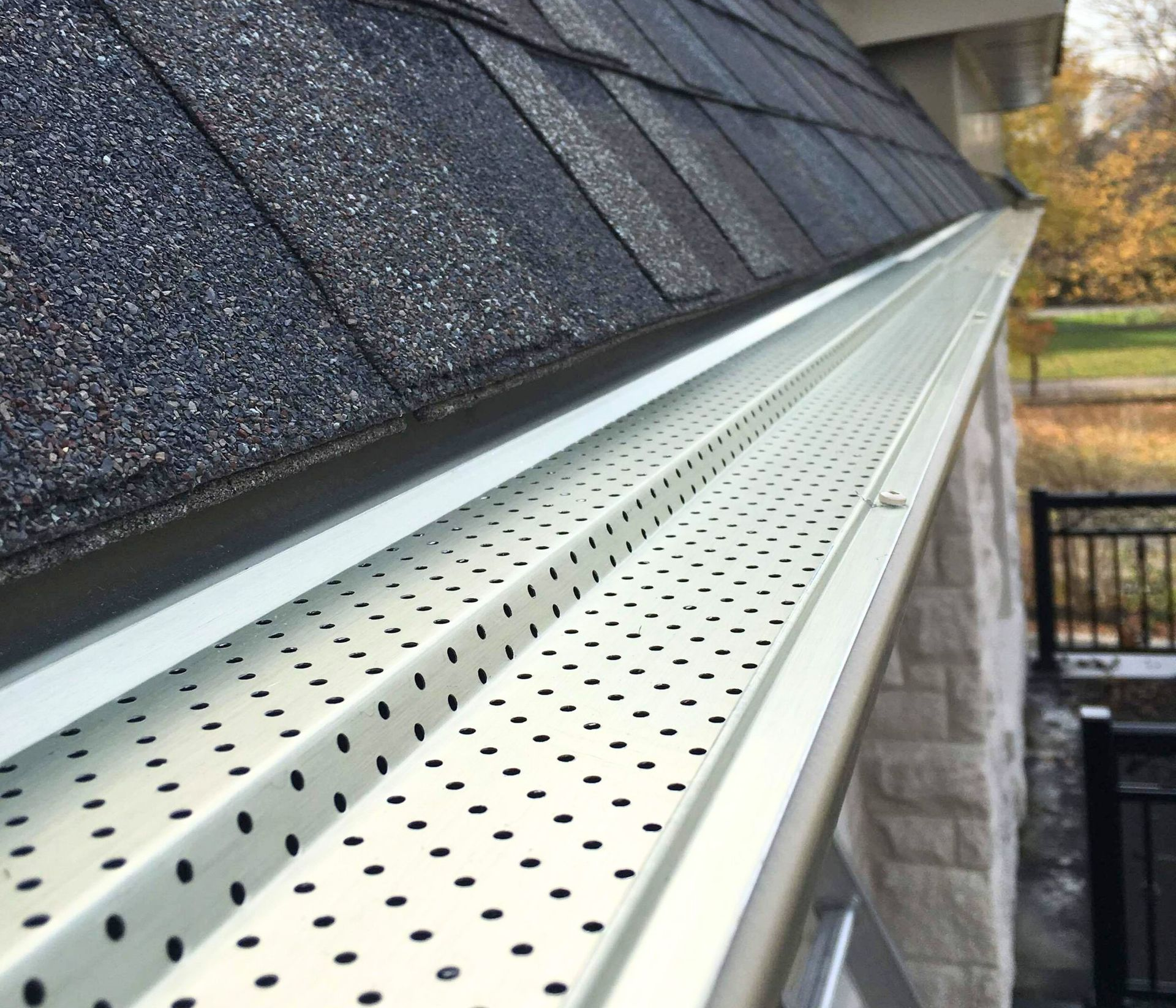 A white metal gutter cover with a perforated pattern installed on a roof with dark asphalt shingles.
