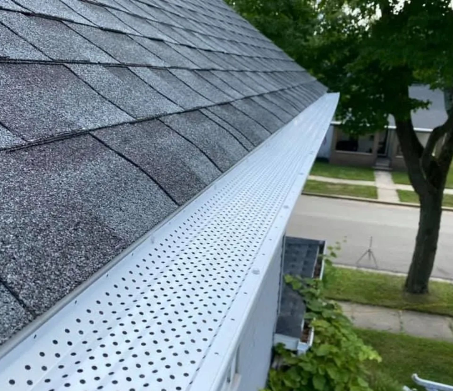 A high-angle view of a gray asphalt shingle roof featuring a white, perforated metal gutter guard installed.