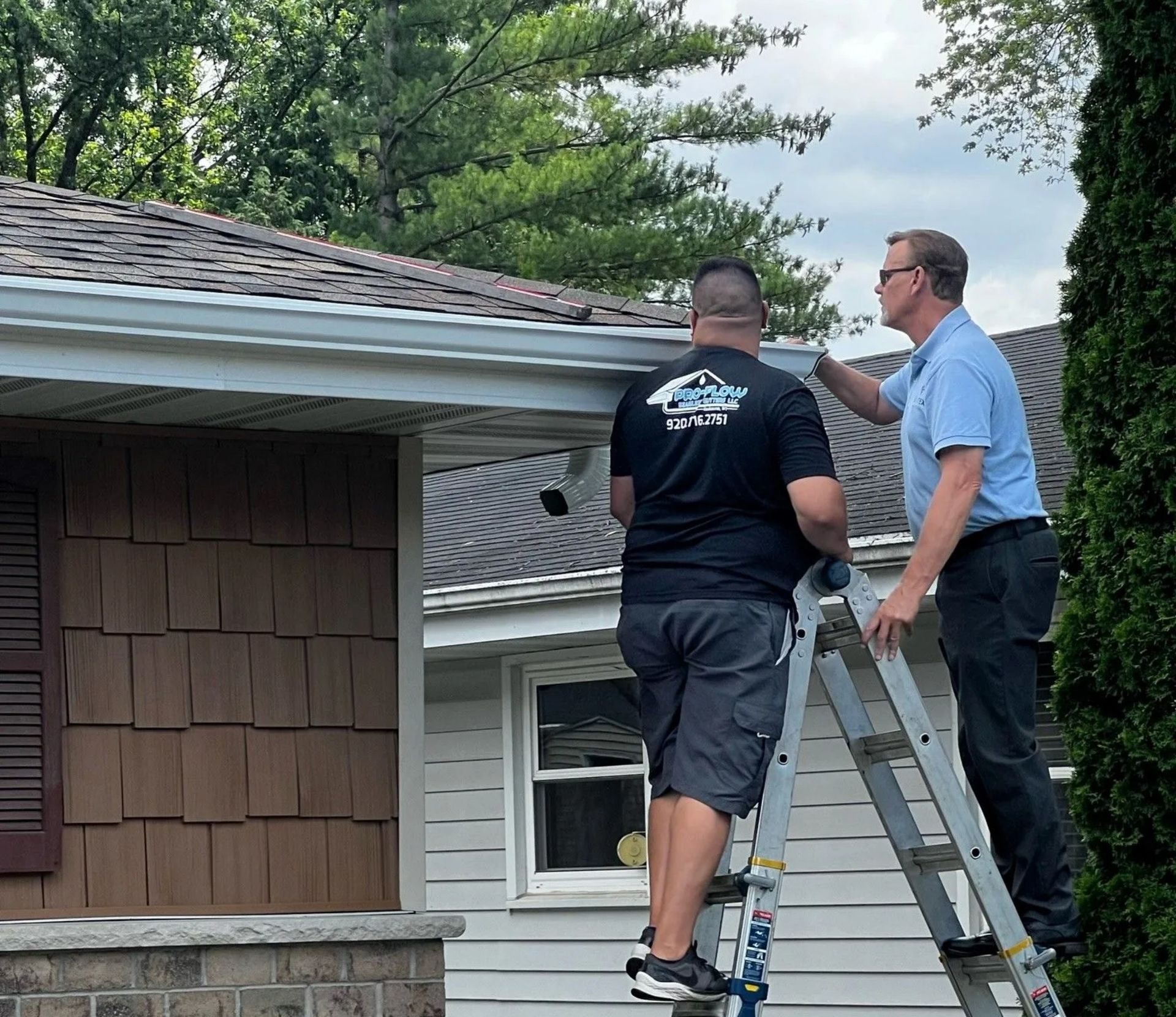 Two people on a ladder inspect the white guttering of a house with brown shake-style siding.