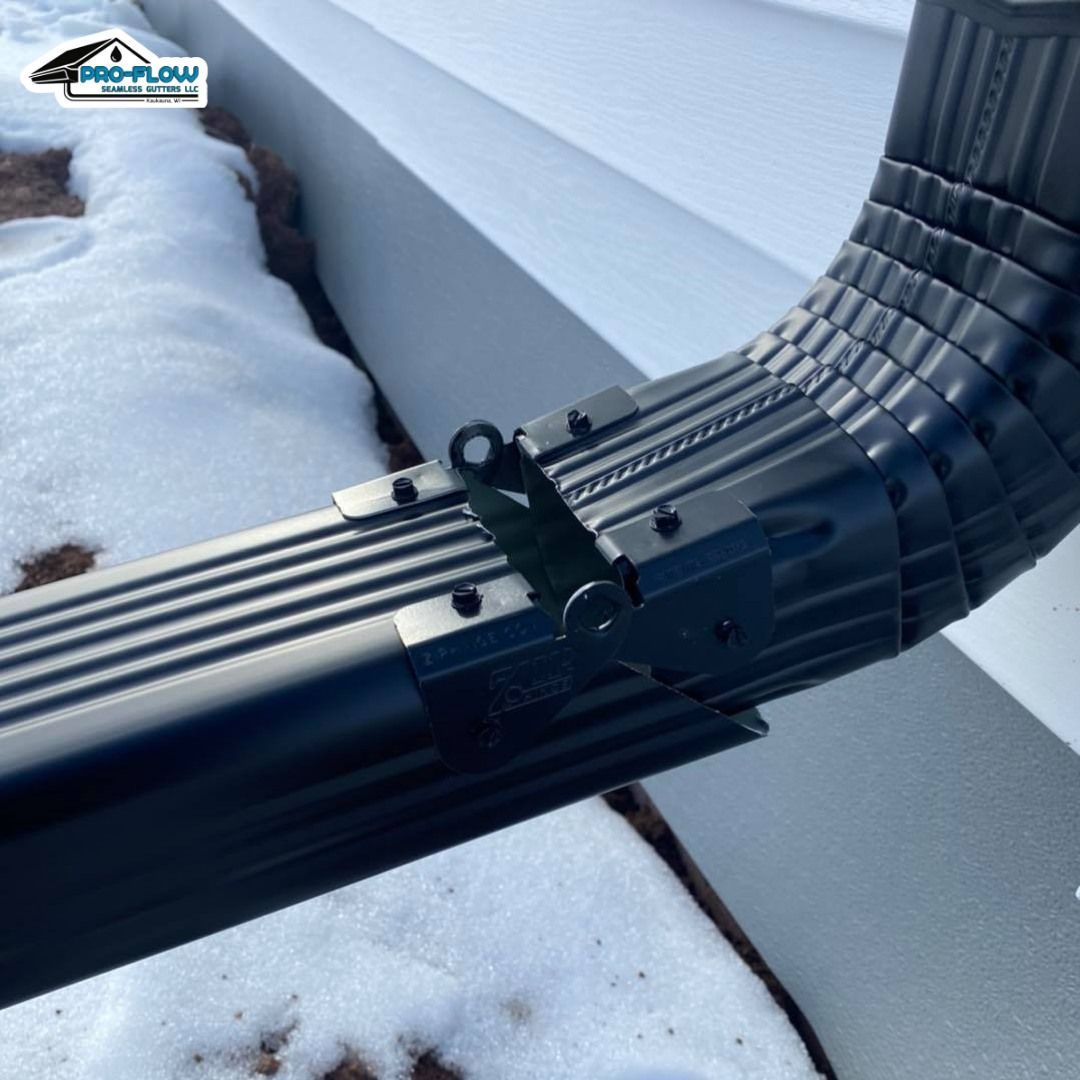 A black downspout extension hinge connected to a gutter downspout against a white house wall with snow on the ground.