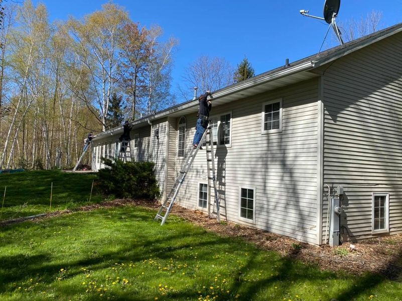 Three people are cleaning gutters on a residential house using ladders on a sunny day.