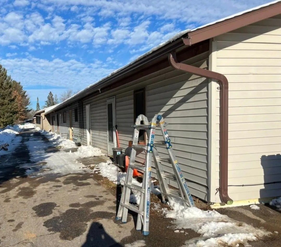 A silver folding ladder stands in front of a beige, single-story building with brown gutters on a sunny, snowy day.