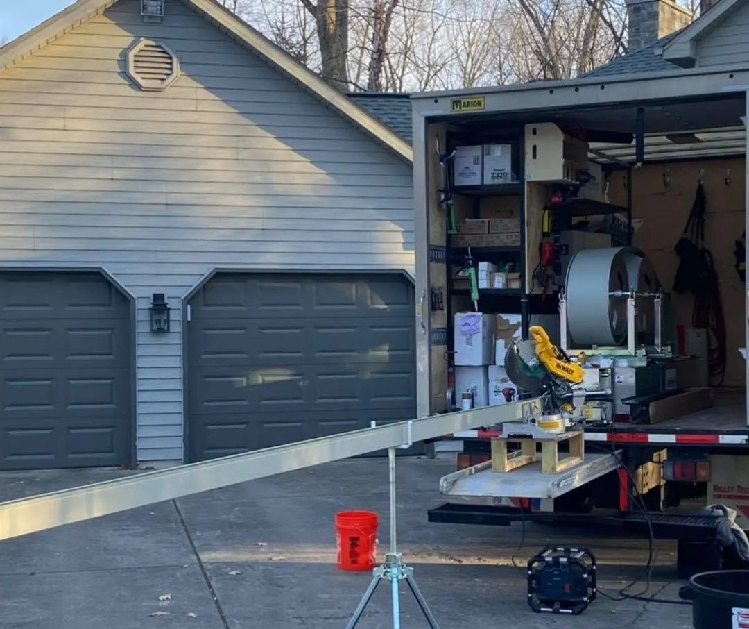 A work trailer parked in a driveway, featuring a miter saw mounted on a long material support stand and a red bucket nearby.