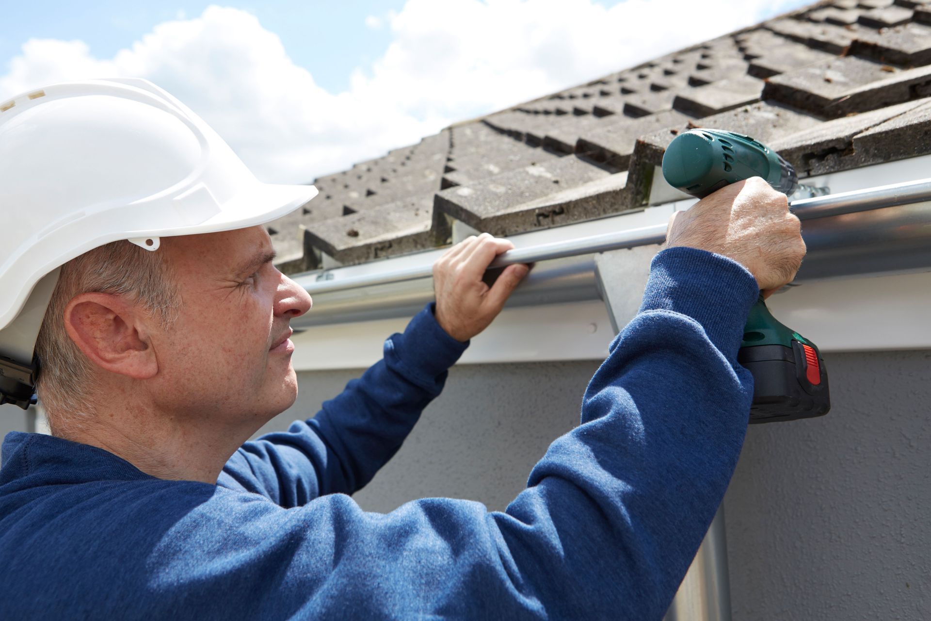 A person in a white hard hat uses a power drill to install a gutter along the edge of a house roof.