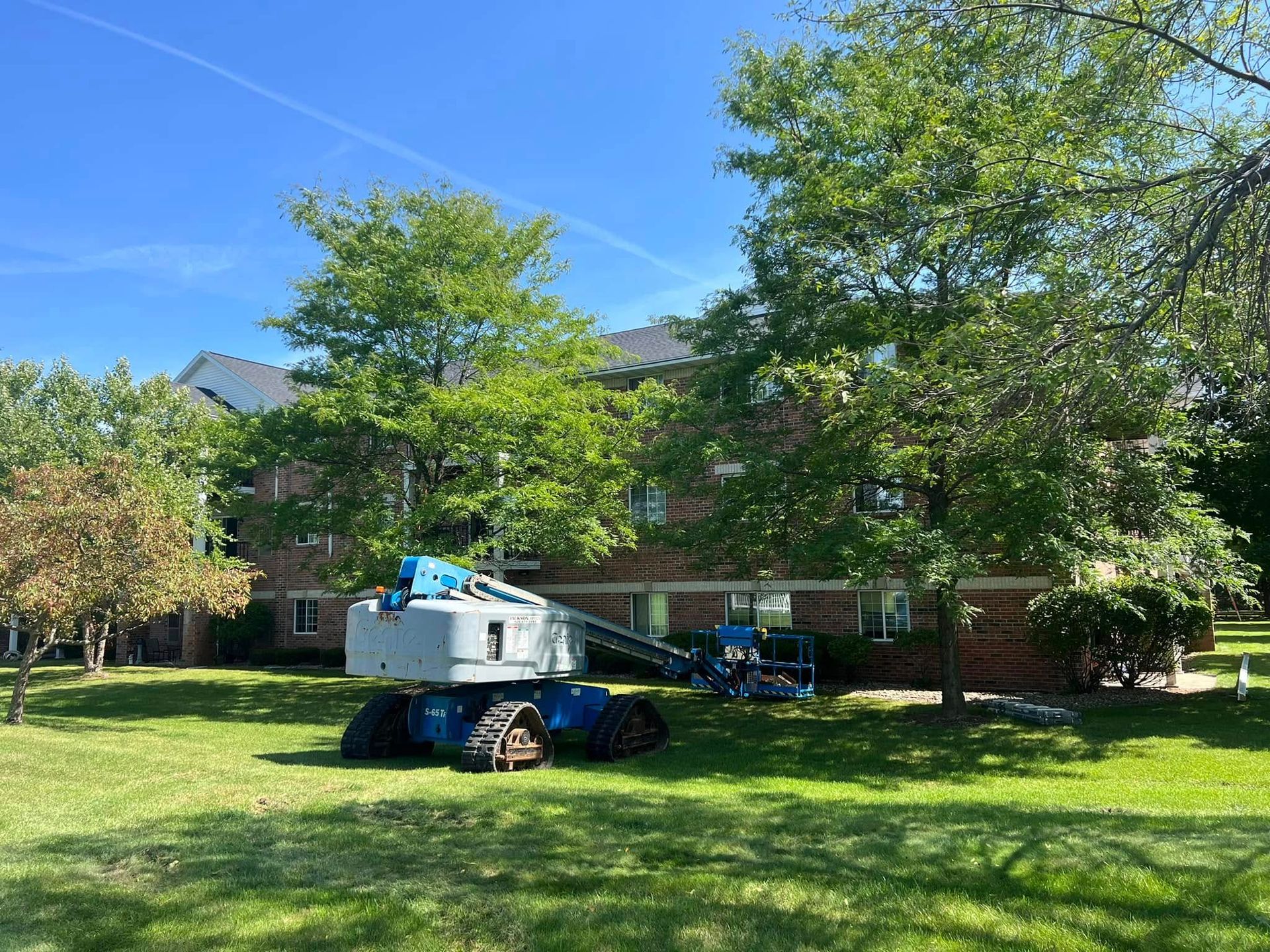 A blue and white tracked boom lift parked on a grassy lawn in front of a brick building surrounded by trees.