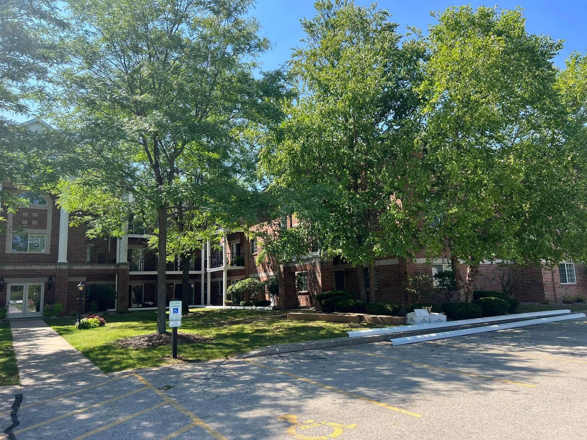 A multi-story brick apartment building set behind a parking lot, surrounded by green trees under a bright blue sky.