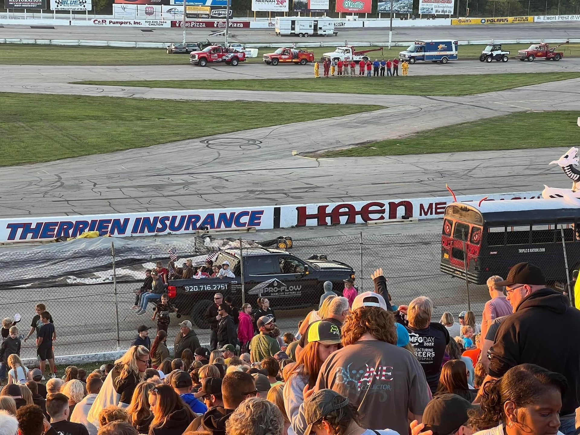 A crowd watches a truck at a demolition derby track near a wall with sponsor signs at an outdoor venue.