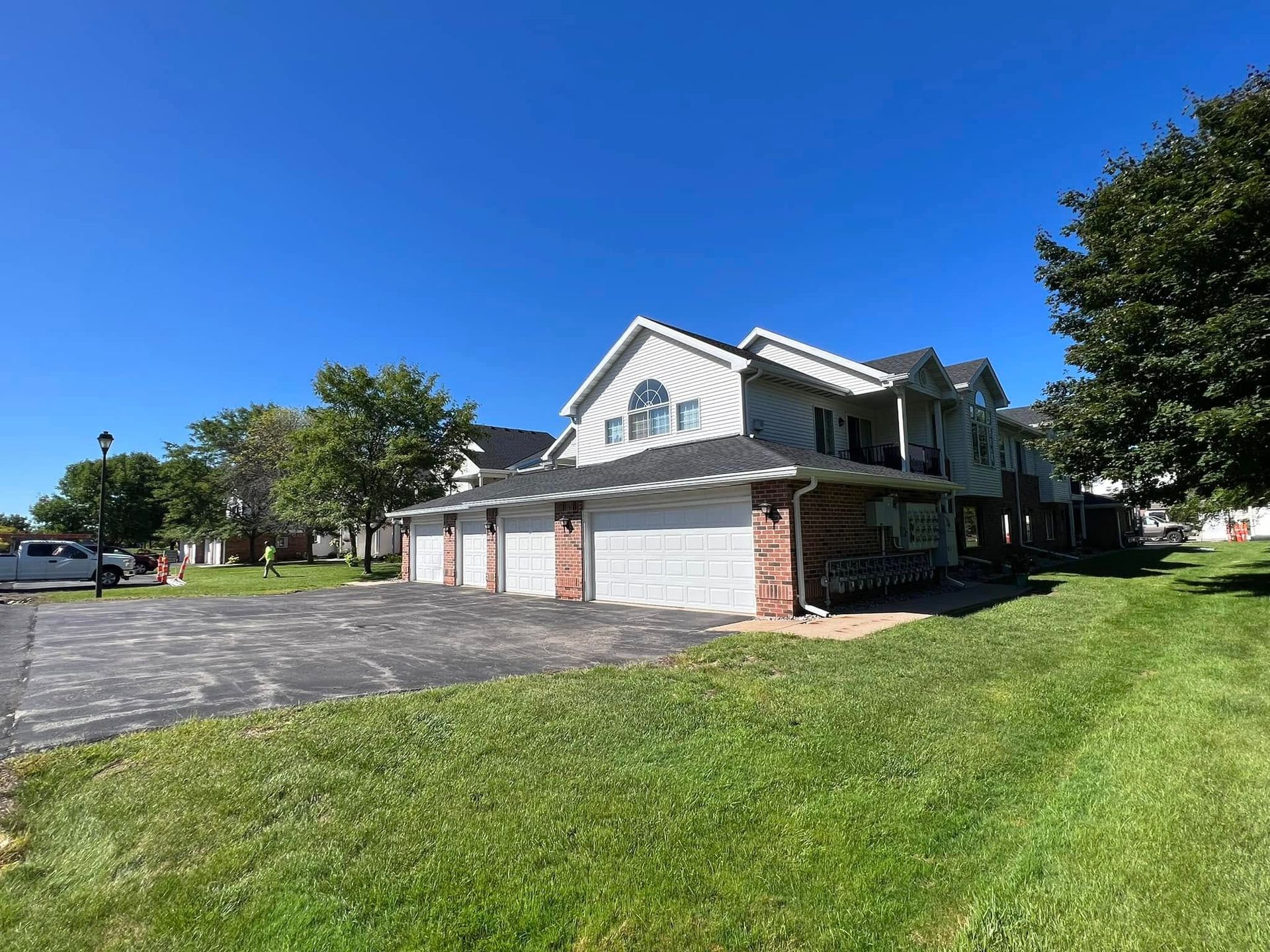 A multi-unit suburban apartment building with gray siding, brick accents, and a long asphalt parking lot under a blue sky.
