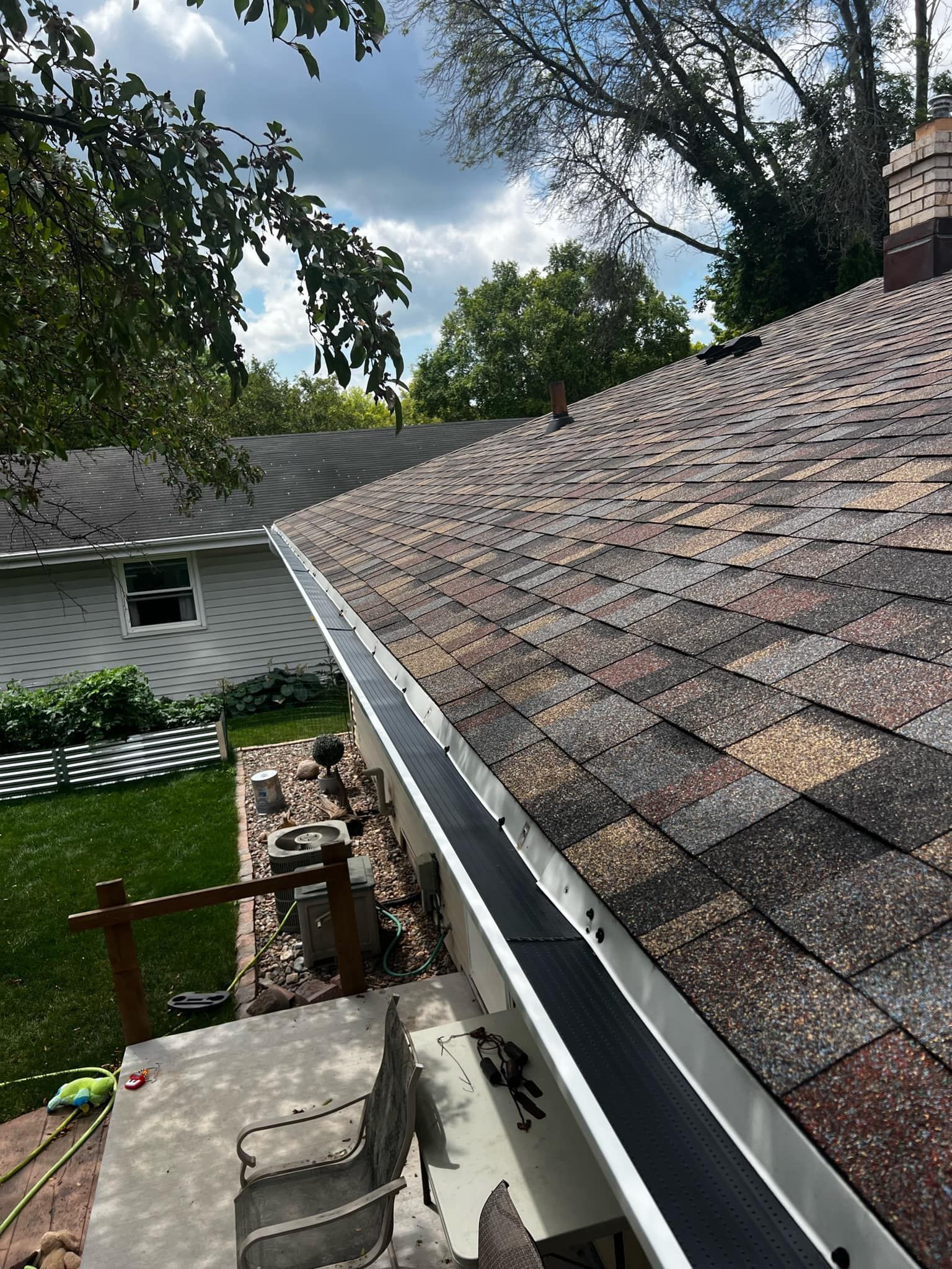 A view looking down from a rooftop with shingled tiles and black gutter guards over a backyard patio area.