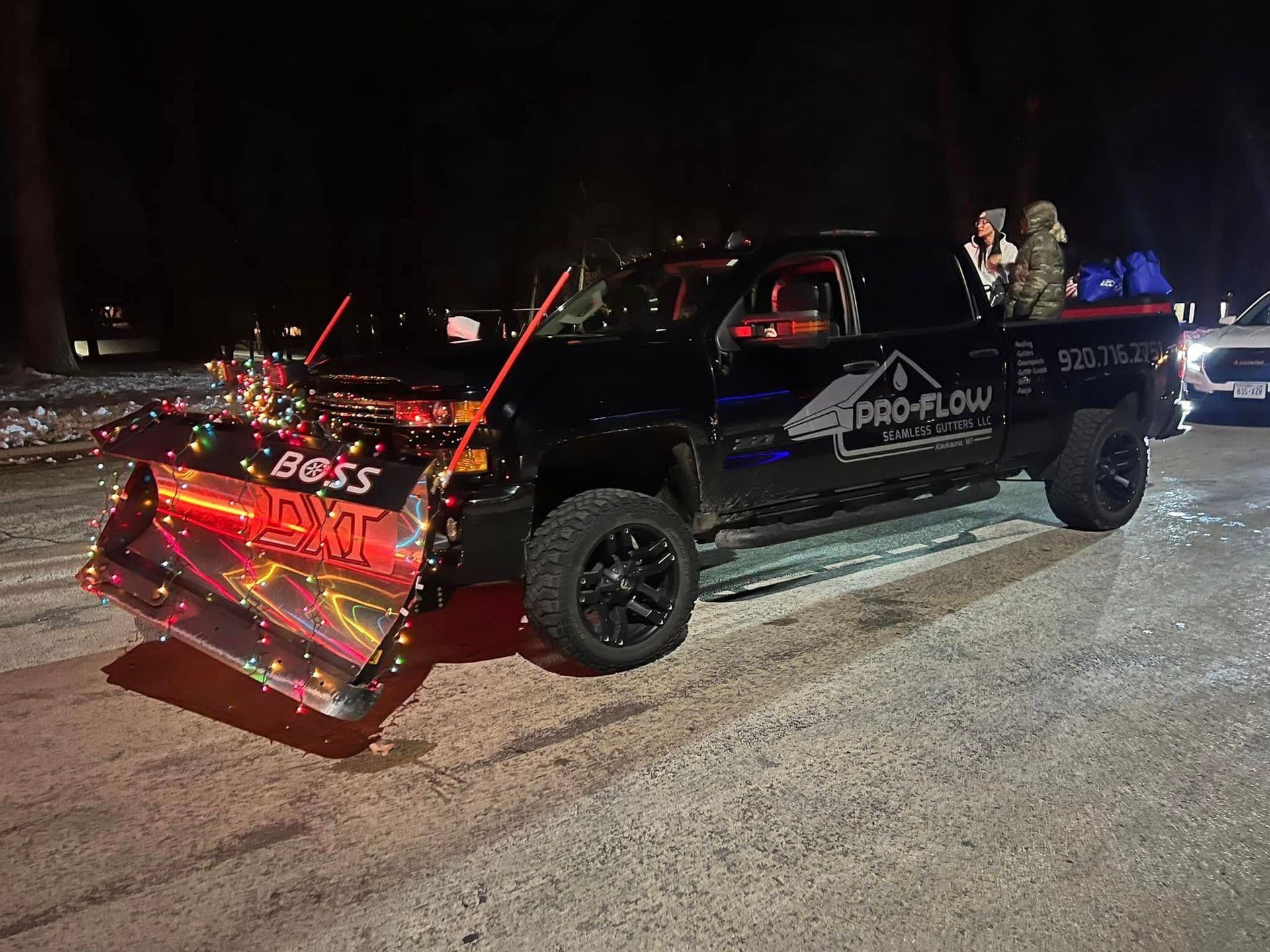 A black pickup truck with a festive, lit-up snow plow attached to the front, parked at night on a snow-covered surface.