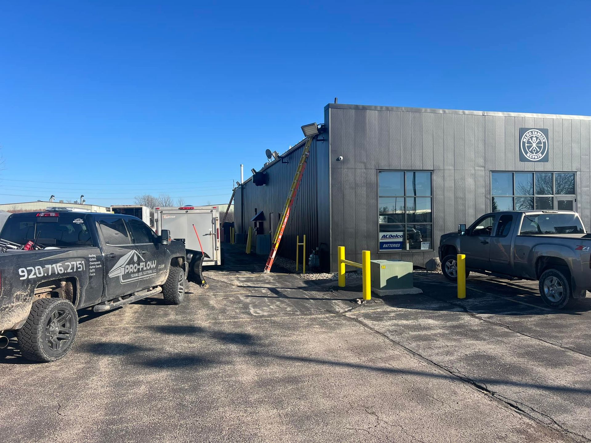 A dark gray metal warehouse building with two pickup trucks and a trailer parked outside on a dirt lot under a blue sky.