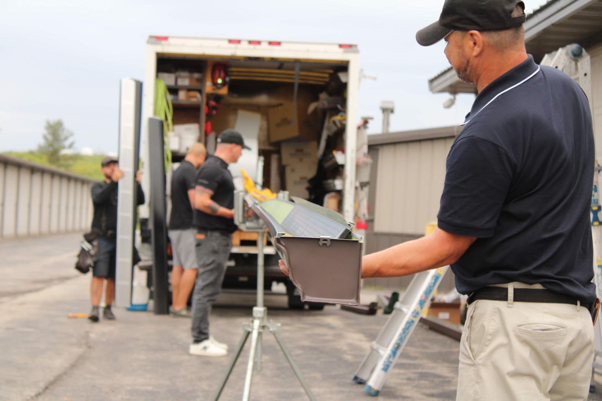 Workers load equipment from a truck at a storage facility, with one foreground worker holding a metal gutter component.