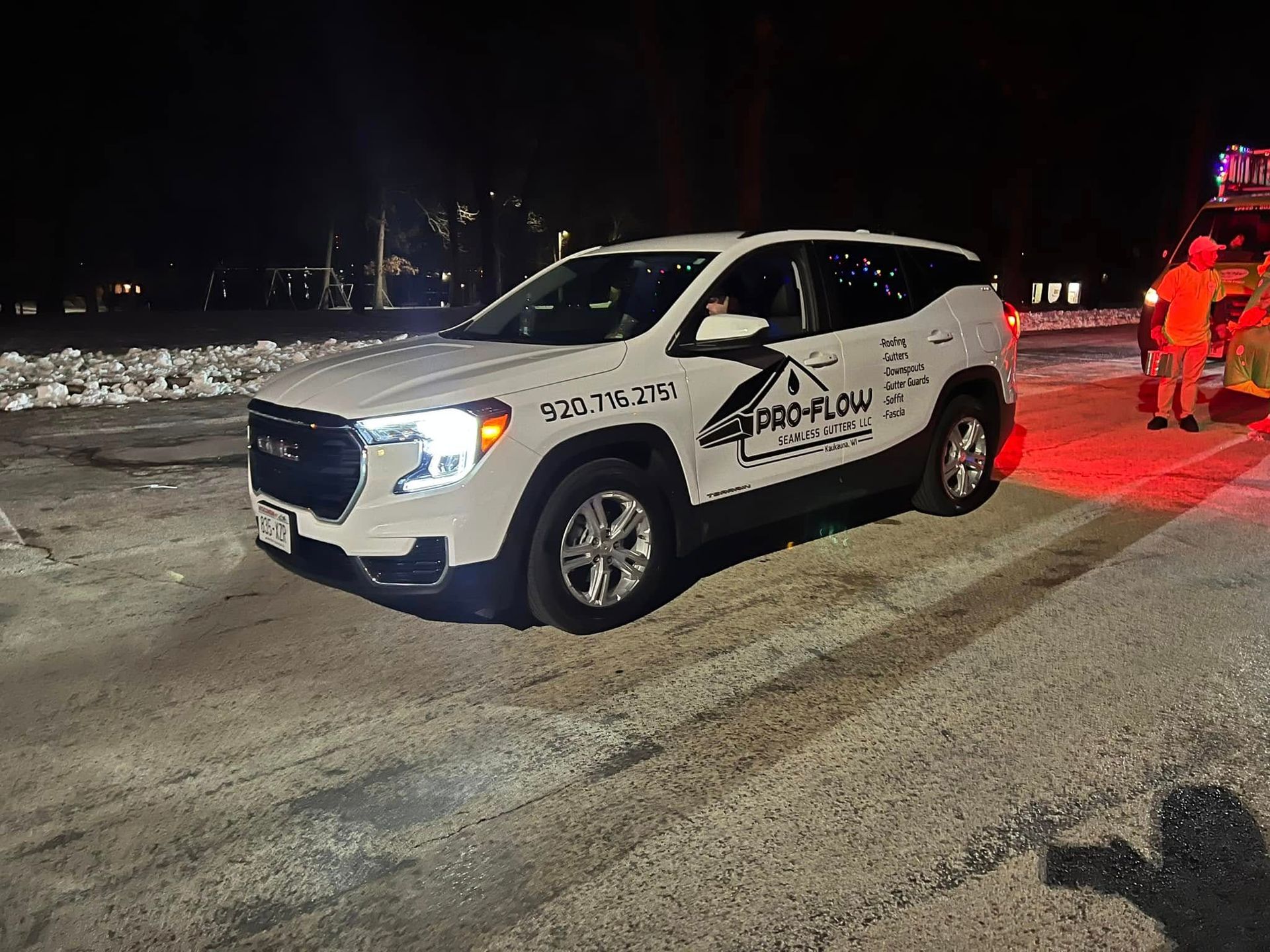 A white GMC SUV with business branding parked on a snowy road at night, with emergency personnel visible in the background.