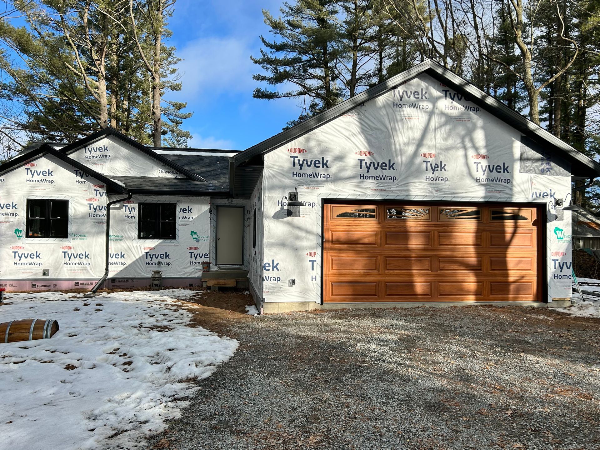 A single-story house under construction, wrapped in white Tyvek, featuring a brown garage door and a gravel driveway.
