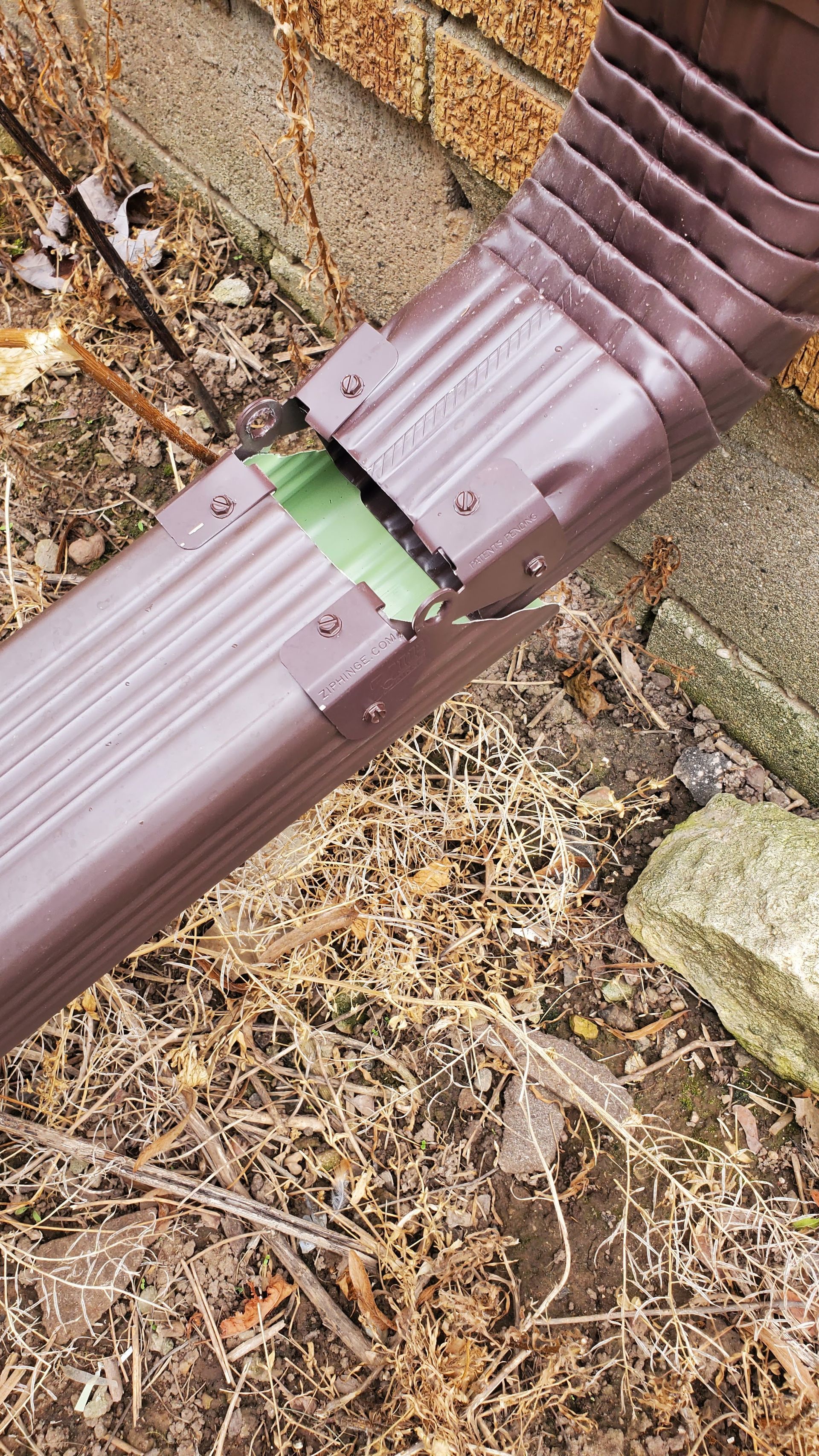 A brown metal gutter downspout is broken and disconnected at the elbow joint near a brick wall and dry, dead foliage.