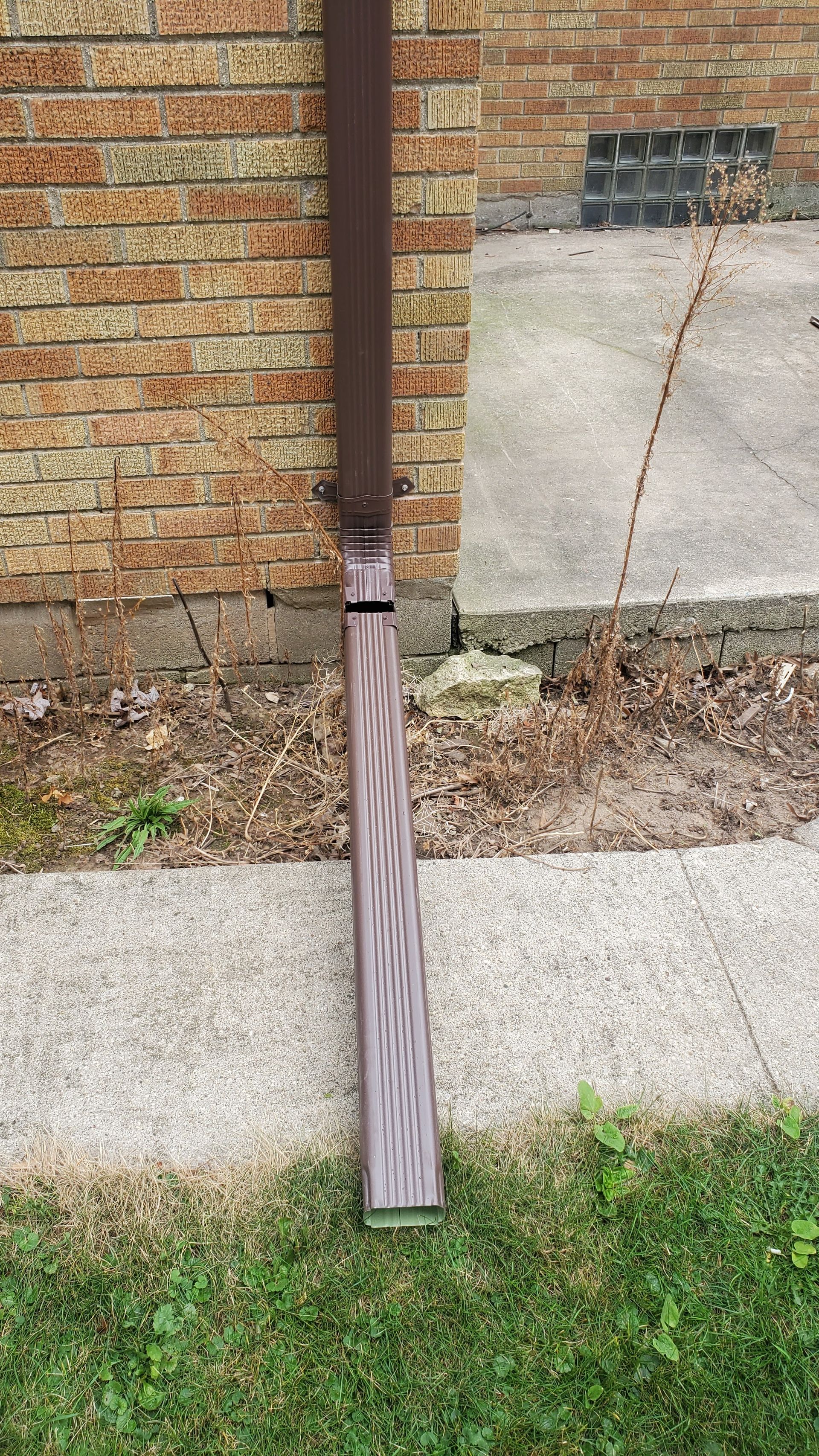 A brown downspout extending from a brick wall onto a concrete walkway next to a grassy area.