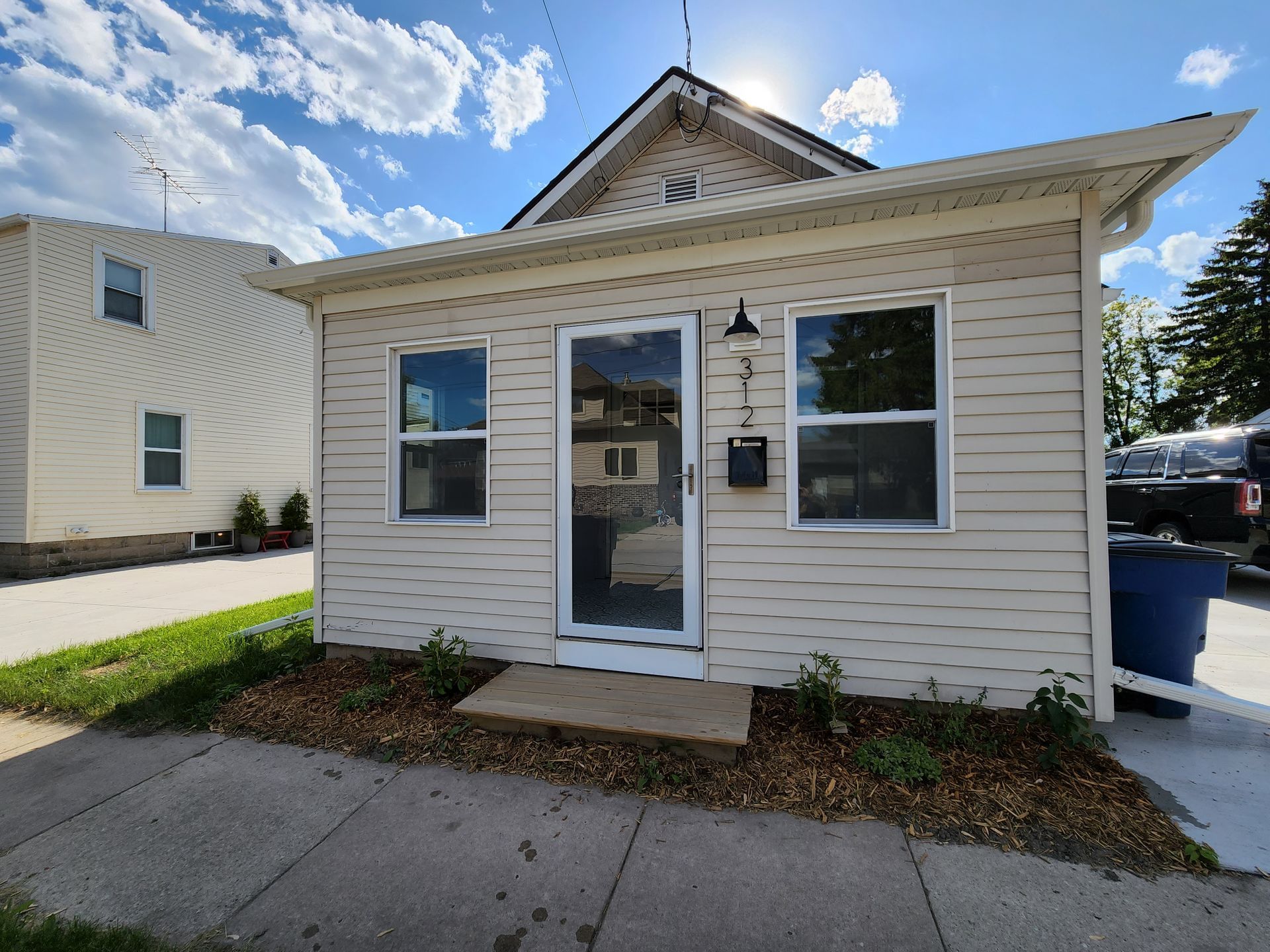 A light-colored, single-story house exterior with white siding, a centered front door, two windows, and a small porch.