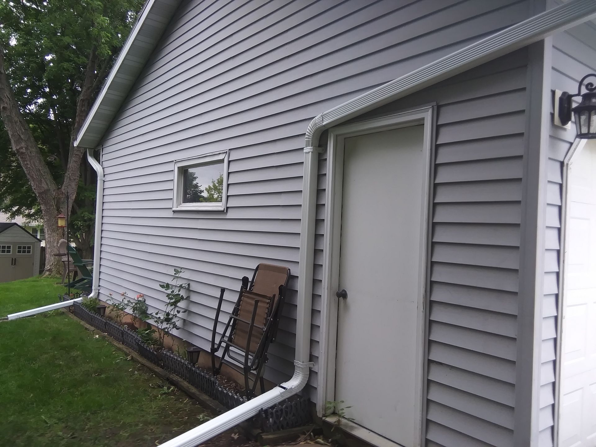 A side view of a house with gray vinyl siding, a small window, a white door, and a white downspout on a lawn.