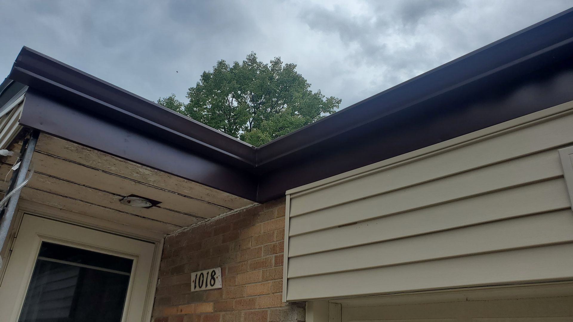 A low-angle view of a house exterior showing dark brown gutters against tan siding, a brick wall, and house number 1815.