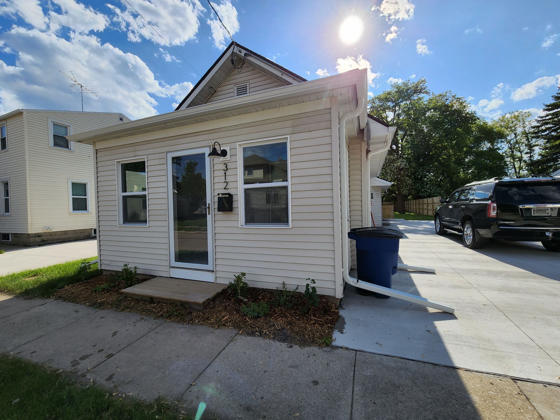 A cream-colored, single-story house with a front door and a driveway with a dark SUV, under a bright, sunny sky.