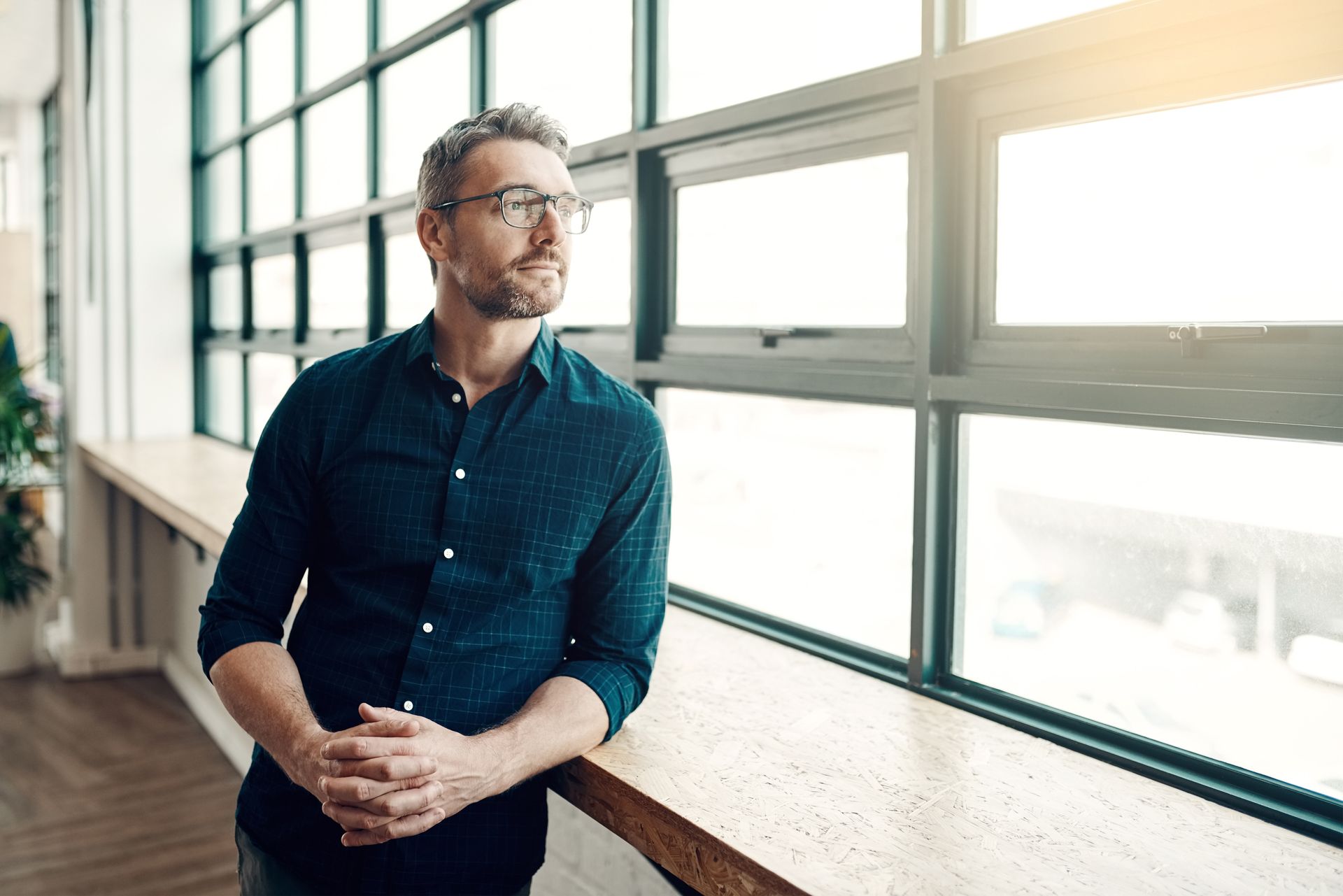 Man clasping hands leaning against a long wooden table looking thoughtfully out of an office window
