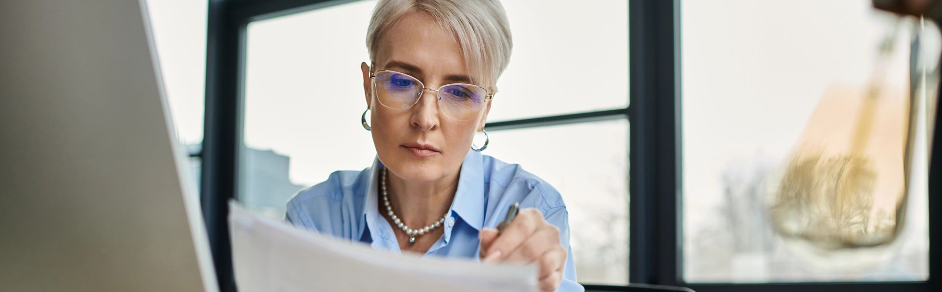 Mature woman with short silver hair wearing glasses and reading paperwork with window in background and part of computer screen in left foreground. 