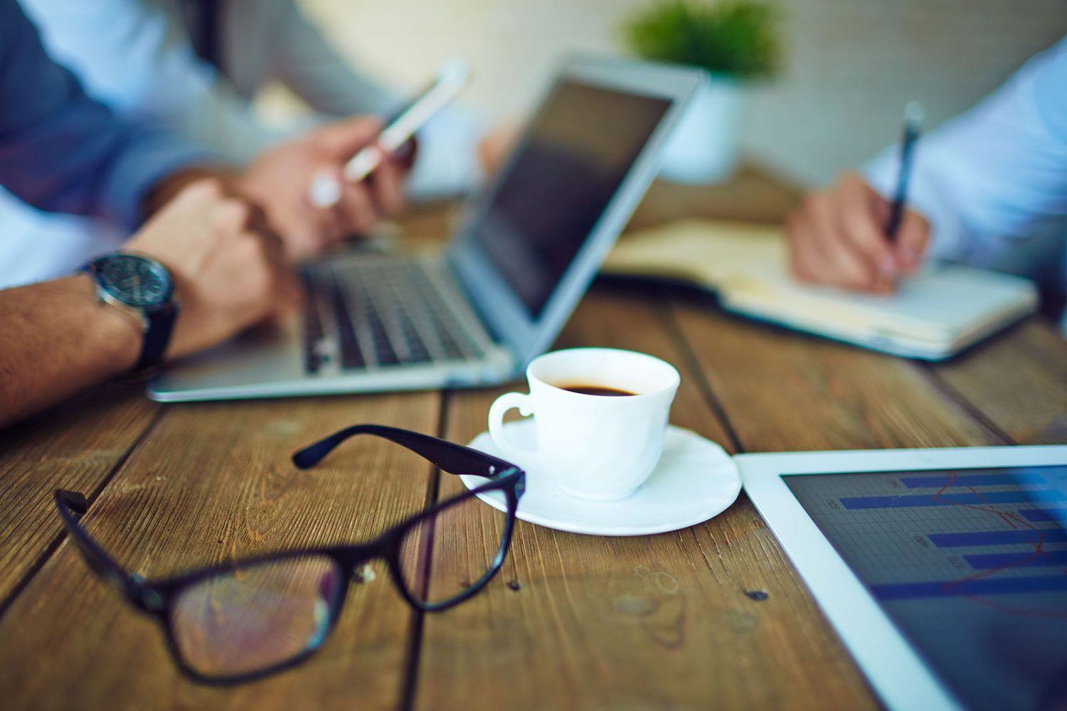 Blurred image of a wooden table with a pair of eyeglasses and a white espresso cup and saucer sitting nearby. Also nearby are a computer tablet, a laptop and what appears to be a man wearing an analog watch holding a cell phone in one hand and the other hand resting on the laptop. To the left is someone writing in a notebook. 
