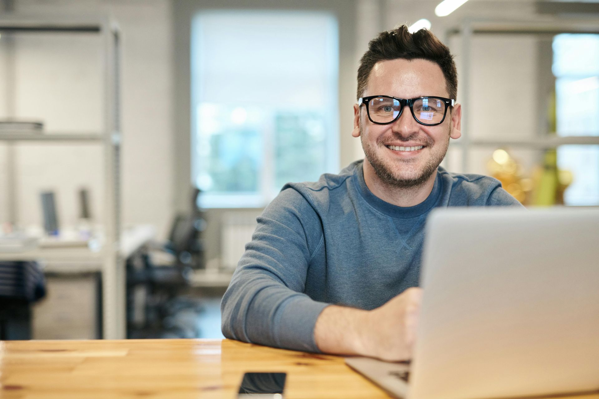 Man at desk with laptop open smiling and wearing glasses. 