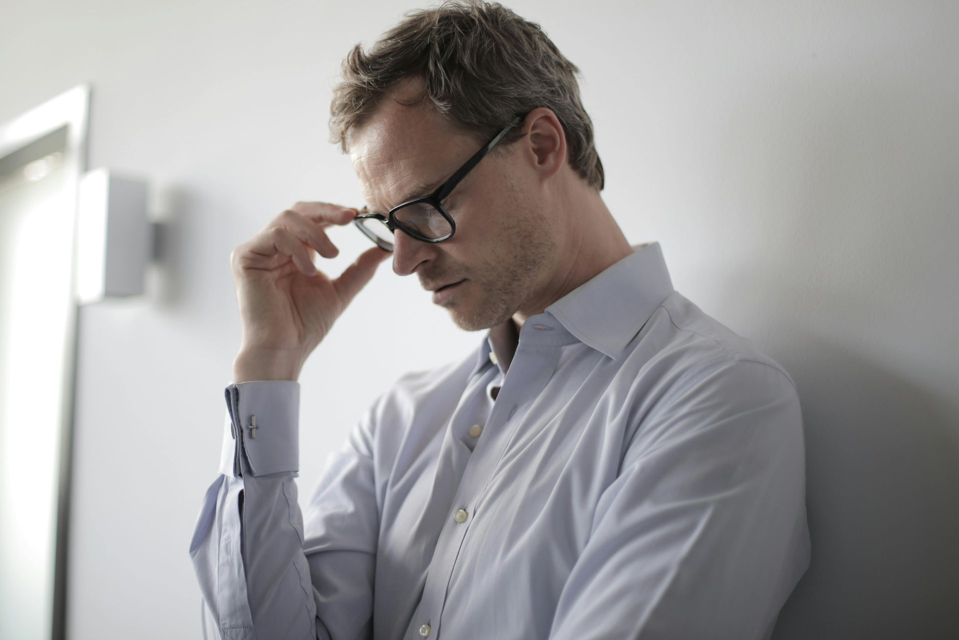 man wearing glasses with right fingers on right edge of glasses looking down feeling frustrated.