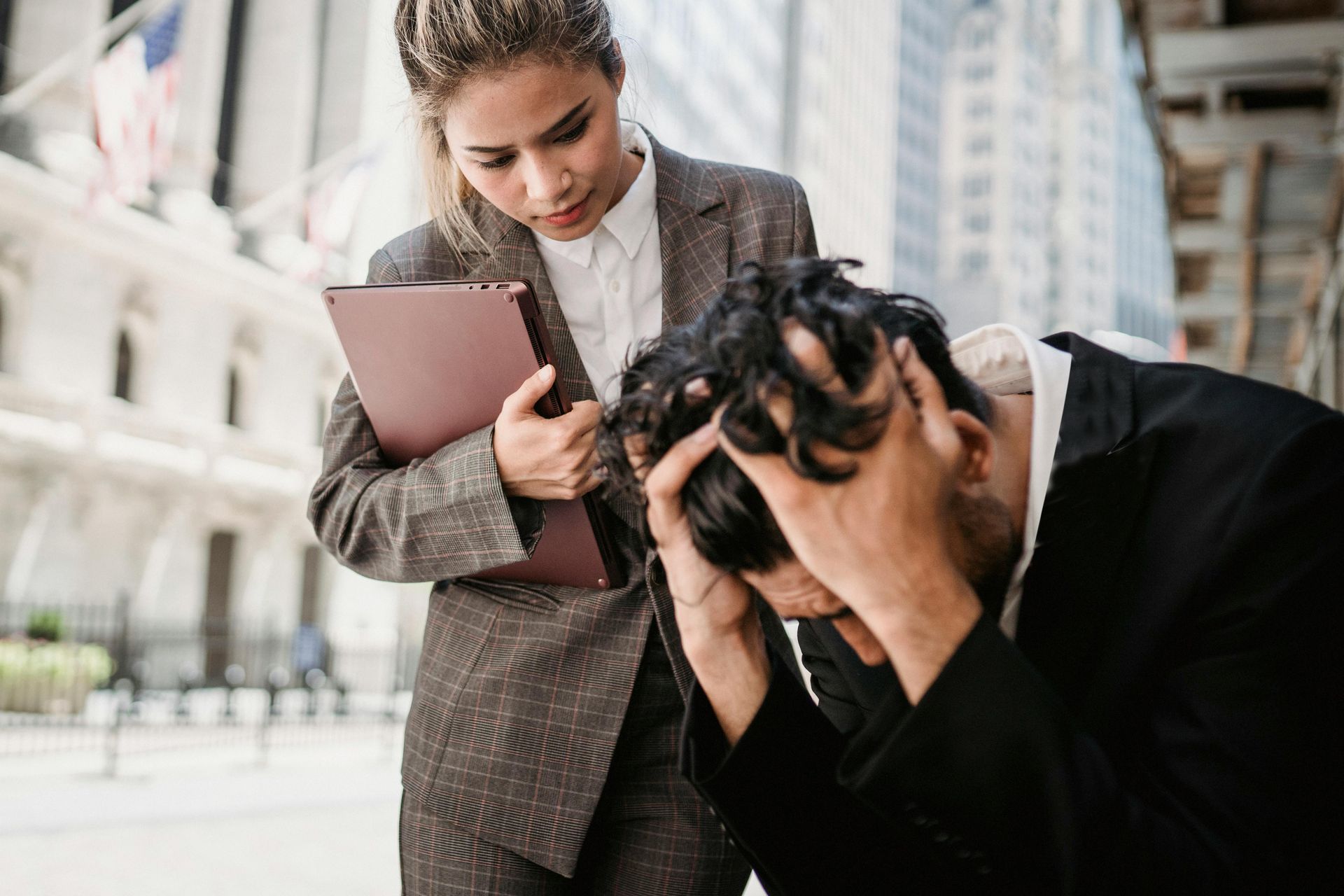 young woman in suit with laptop looking concerned over man in suit sitting with hands in hair seemin