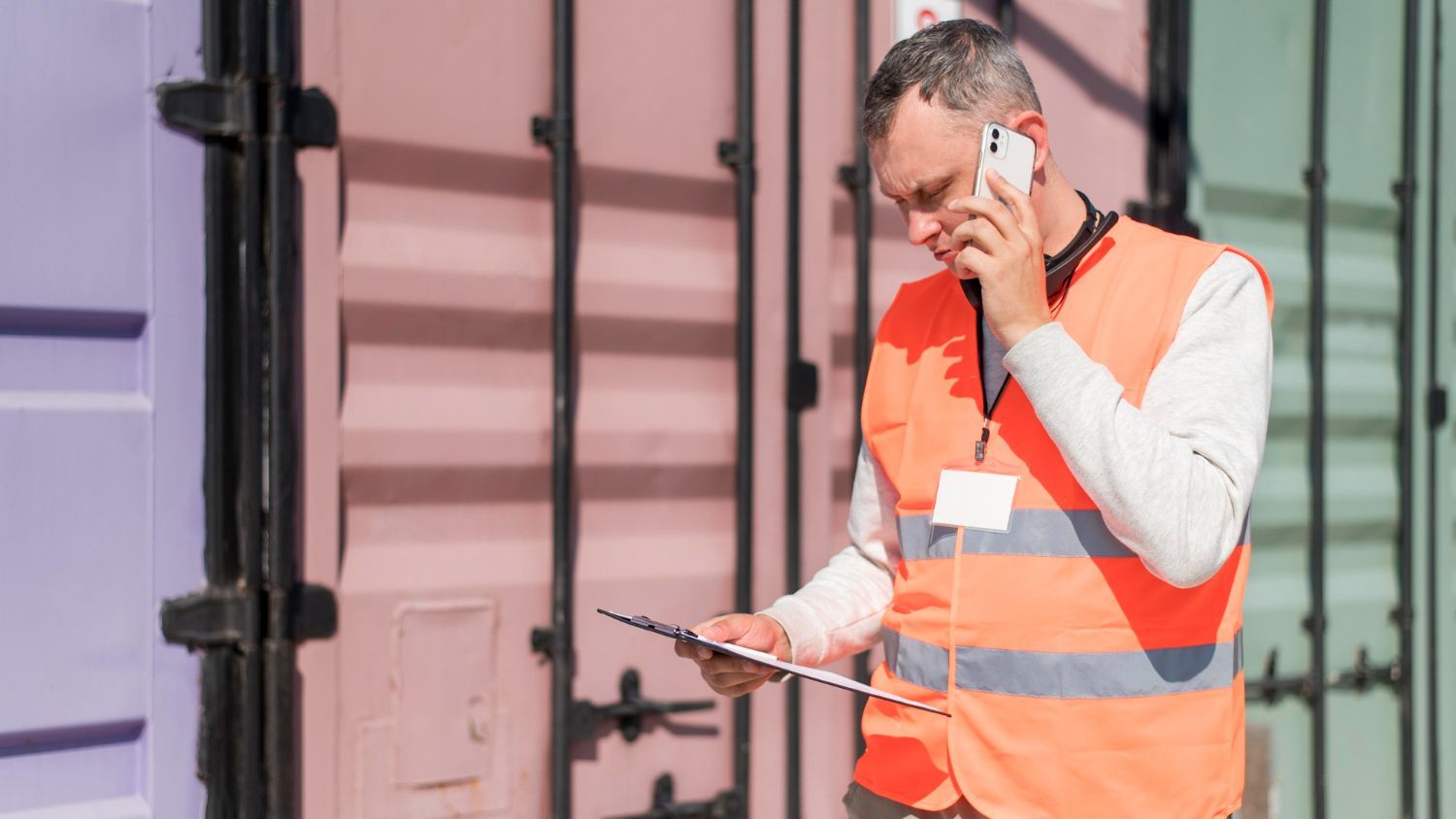 A man in an orange vest is talking on a cell phone while looking at a clipboard.