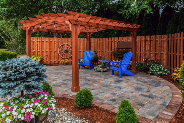 Wooden pergola over a patio with blue chairs. Brown fence surrounds the space, with flowers and greenery.