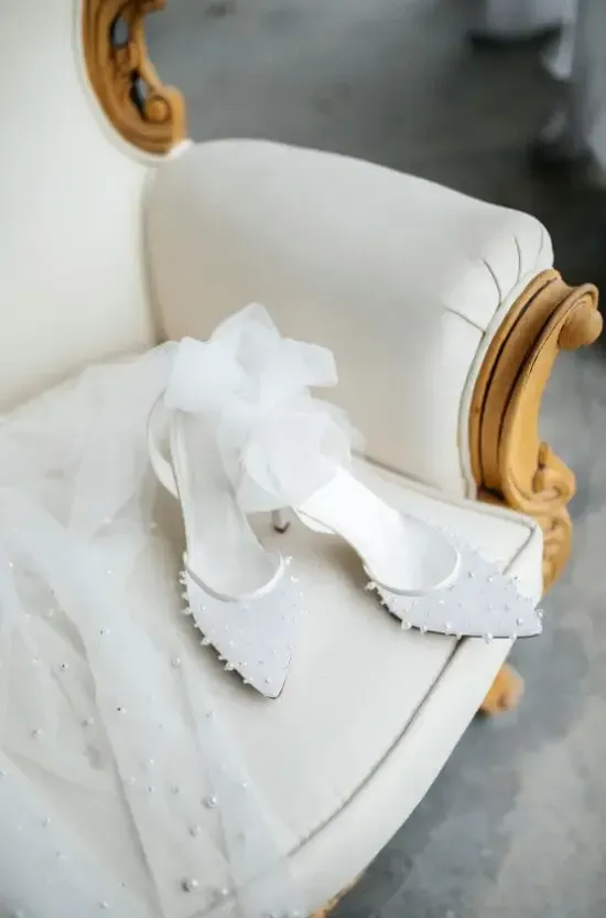 White bridal shoes with a bow, resting on a cream-colored ornate chair.