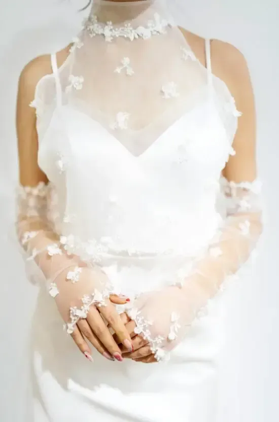 Woman in a white wedding dress with a high-neck sheer overlay and floral appliqués, hands clasped.