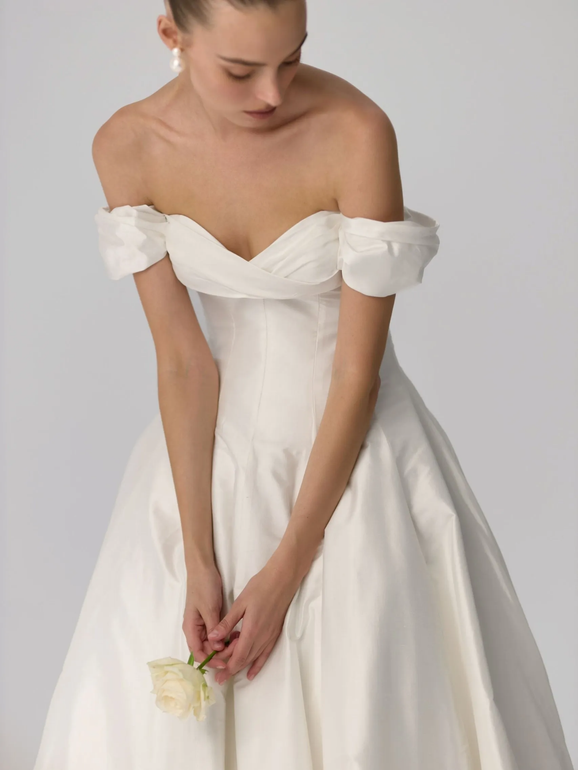 Woman in a white off-the-shoulder wedding dress, holding a white flower, looking down.