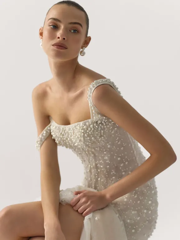 Woman in a beaded, off-the-shoulder gown, sitting, looking toward the viewer; light background.