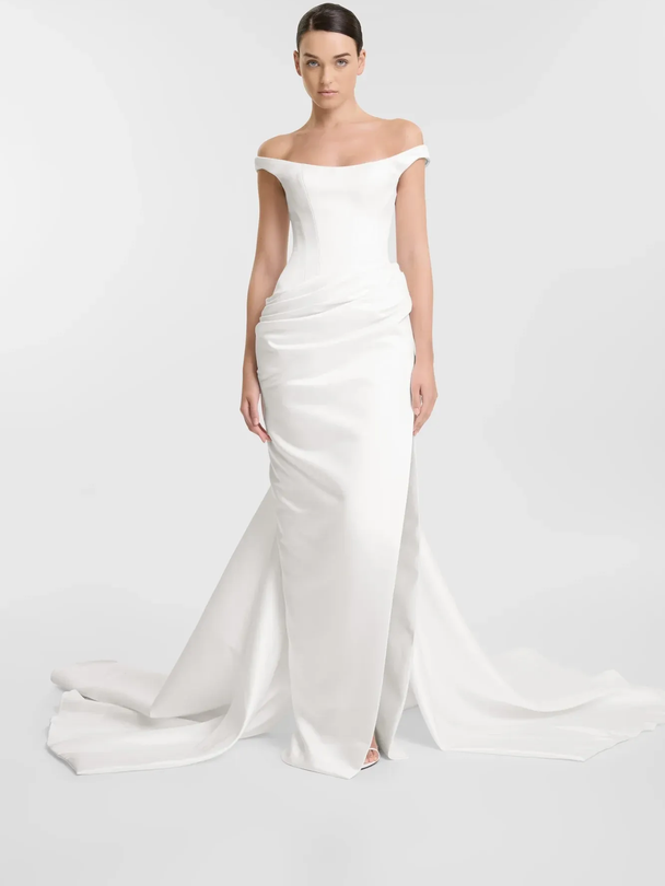 Woman in white off-the-shoulder wedding gown, standing in a studio with a long train.